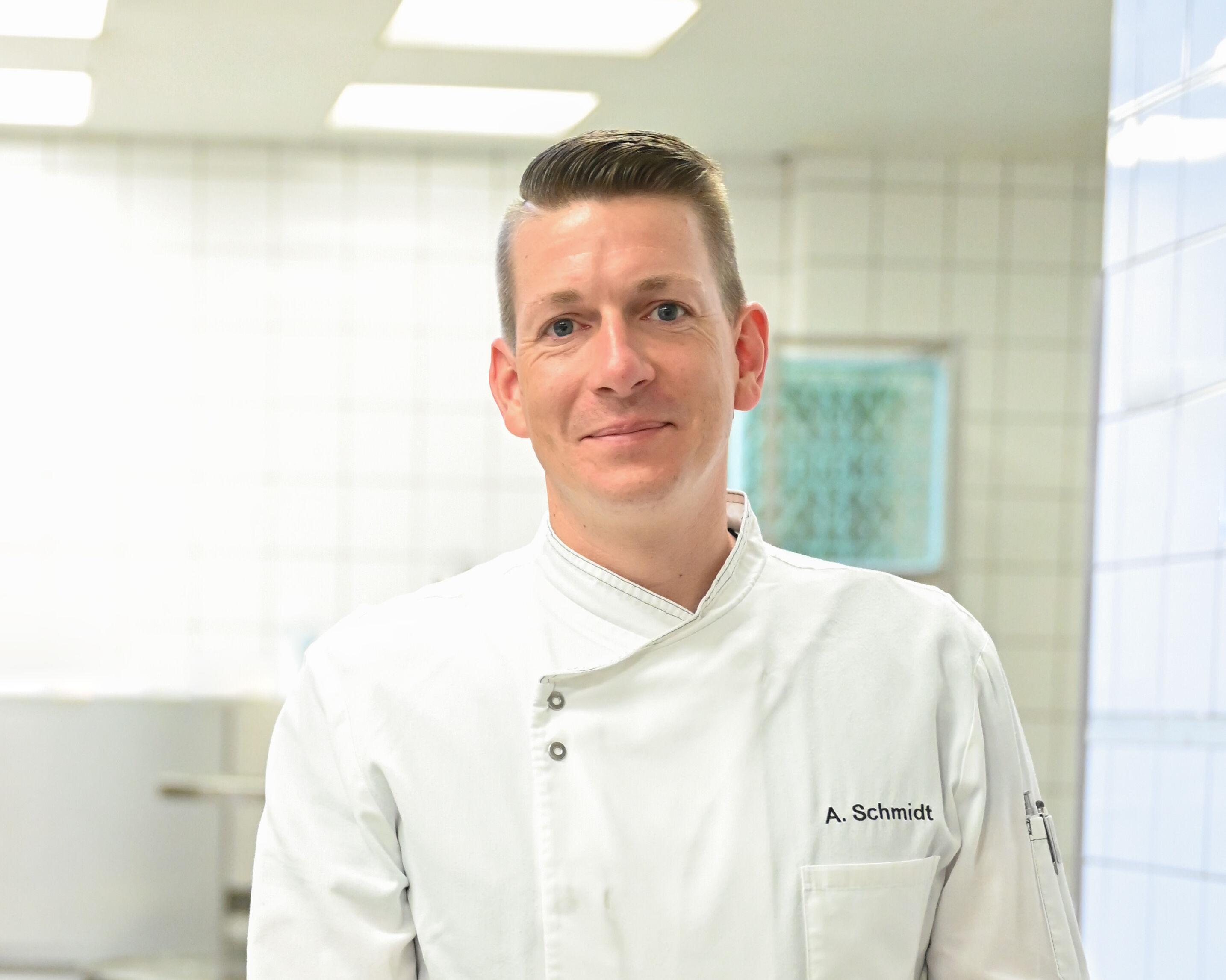 Mann mittleren Alters in Kochschürze steht lächelnd in einer Großküche.