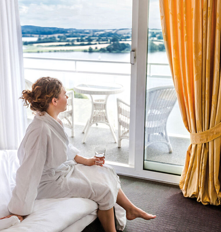 Eine junge, lockige Frau im Bademantel sitzt auf der Kante Ihres Bettes und blickgt auf den Segeberger See.