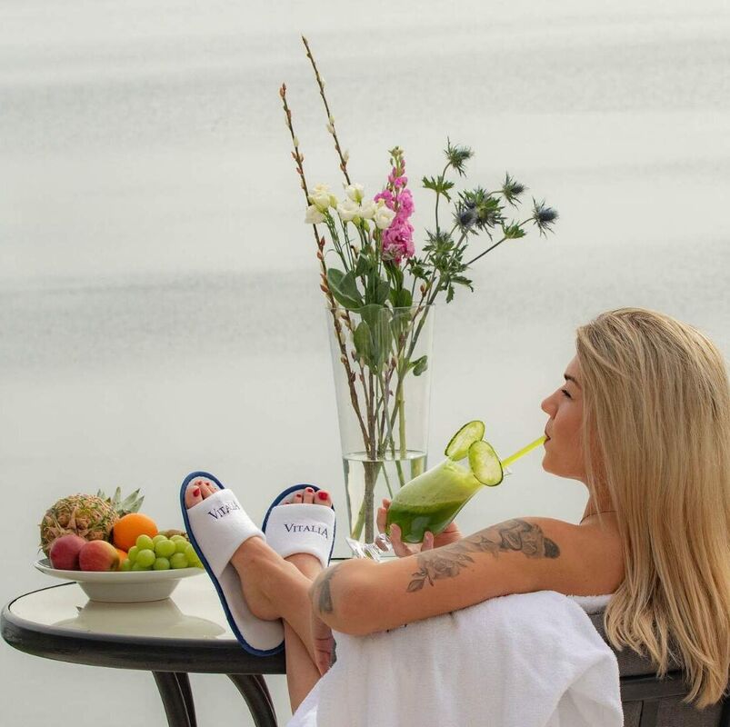Blondine sitzt in Bademantel an gedecktem Tisch mit Wellnessdrink am See